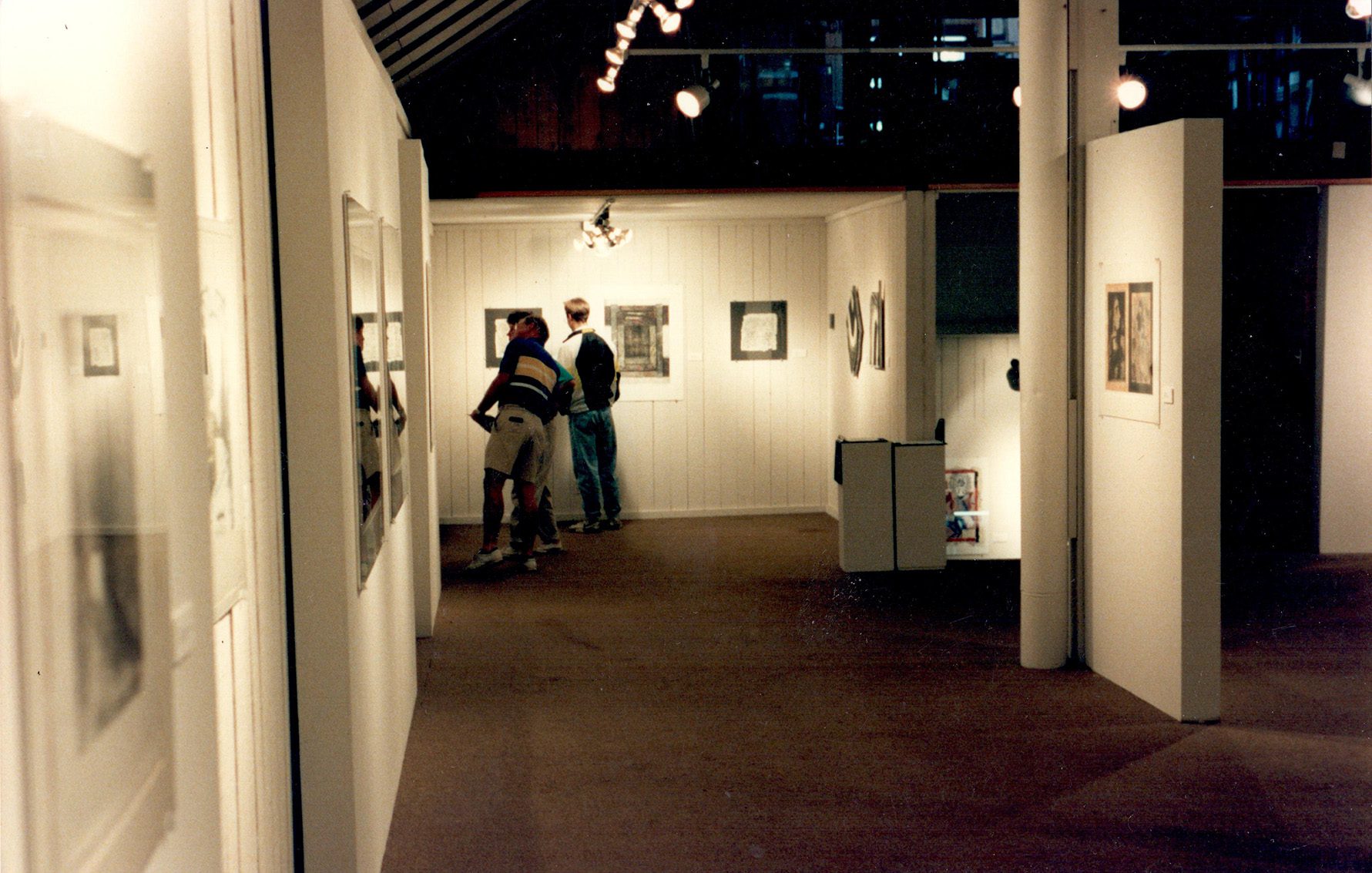 Installation view of Exchange, The Bush Barnes Art Centre, 1992. There are two viewers looking at work in a white paneled gallery.